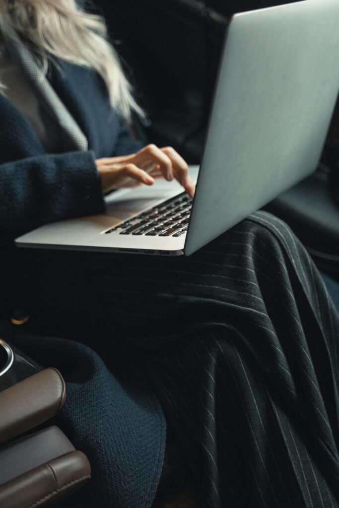 Woman in a blue coat typing on a laptop while sitting in an office setting.